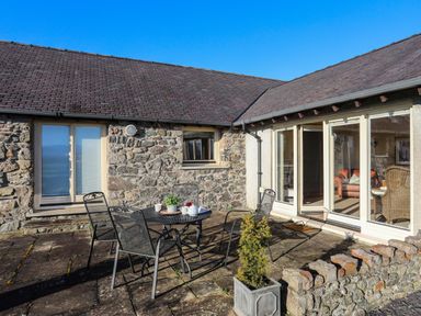 An outdoor patio with a round metal table and four metal chairs next to stone walls and windows at Cosy Barn in Llangaffo