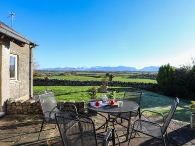 A patio with metal table and chairs overlooking a stone wall and green fields with mountains in the distance at Cosy Barn in Llangaffo