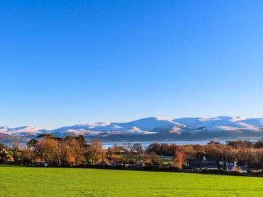 A green field with trees and houses in front of snow covered mountains under a clear blue sky at Cosy Barn in Llangaffo