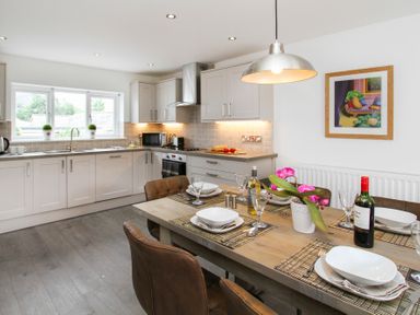 A kitchen with white cabinets and gray countertops and a wooden dining table with chairs set for meals and decorated with a plant at Cuckooland in Borth-Y-Gest
