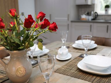 A dining table set with white plates bowls wine glasses and a vase with red roses in a kitchen at Cuckooland in Borth-Y-Gest