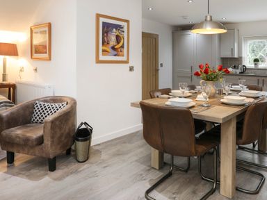 A dining area with a wooden table set with plates and glasses and leather chairs next to a kitchen and a leather armchair with a cushion at Cuckooland in Borth-Y-Gest