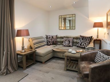 A living room with a sectional sofa, patterned pillows, two table lamps on side tables, a leather armchair, and a small table with tea cups and pastries at Cuckooland in Borth-Y-Gest