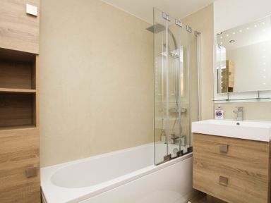 A bathroom with a bathtub and glass shower screen a sink with a wooden cabinet and a mirror at Cuckooland in Borth-Y-Gest