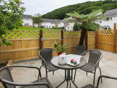 An outdoor patio with a round table and four chairs a teapot cupcakes and a plant on the table with a wooden fence and houses in the background at Cuckooland in Borth-Y-Gest