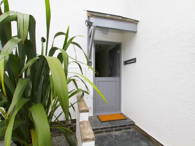 An open gray door with an orange doormat in a white wall entrance with large green plants at Cuckooland in Borth-Y-Gest