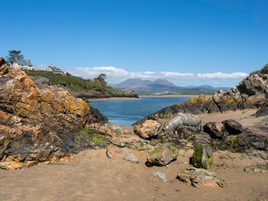 A sandy beach with large rocks and boulders by the water with houses on a hillside in the background at Cuckooland in Borth-Y-Gest