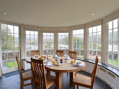 A dining area with a round wooden table and six chairs set for breakfast in a room with large windows at Dinas Noddfa in Nant Gwynant