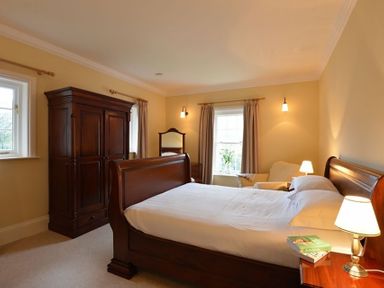 A bedroom with a wooden bed a wardrobe a chair a bedside table with lamps and windows with curtains at Dinas Noddfa in Nant Gwynant