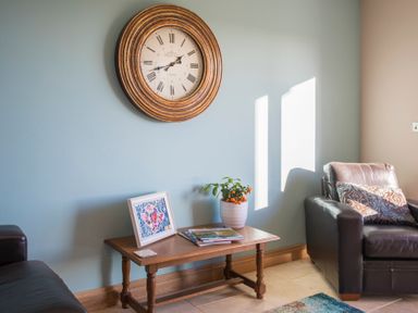 A living room with a wooden table holding a framed picture plant and magazines with a large round clock on the wall and two leather chairs at Dol Fraint in Star