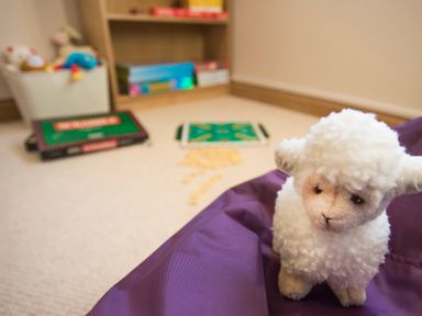 A stuffed lamb toy on a purple bean bag in a playroom with a basket of toys and board games on a carpeted floor at Dol Fraint in Star