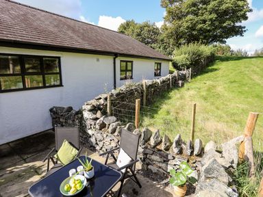 A patio with chairs and a table next to a stone wall and grassy field at Bryn Eira Tack Room in Llanfair PG