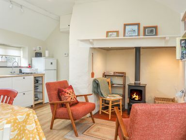 A living area with two red chairs a wood burning stove a small wooden chair with a blanket a kitchenette and a table with a yellow tablecloth at Ffrwd Cottage in Bodorgan