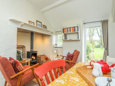 A sitting room with two armchairs by a wood stove and a table with a red teapot near a door to a garden at Ffrwd Cottage in Bodorgan