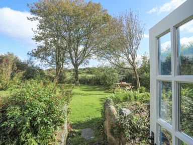 An open door leading to a garden with trees bushes grass and a picnic table at Ffrwd Cottage in Bodorgan
