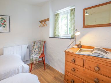 A bedroom with a wooden dresser a mirror a chair with a blanket and a window with green curtains at Ffrwd Cottage in Bodorgan