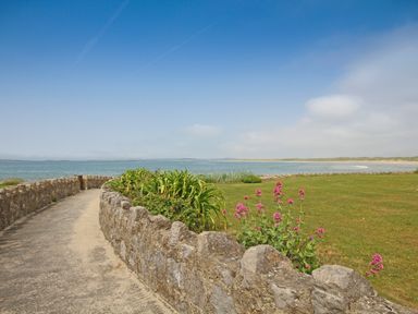 A stone pathway beside a grassy area with flowers and ocean in the background at Flip Flops in Rhosneigr