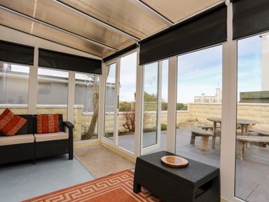 A sunroom with a black wicker loveseat with cushions and orange pillows a black wicker table with a wooden tray and glass doors leading to a patio with a wooden picnic table at Flip Flops in Rhosneigr