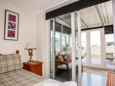 A bedroom with a bed a nightstand with a lamp and a glass door leading to a sunroom with chairs and pillows at Flip Flops in Rhosneigr