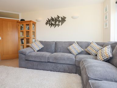A living room with a grey sectional sofa with patterned cushions a wooden cabinet a door and a metal fish wall decoration at Flip Flops in Rhosneigr