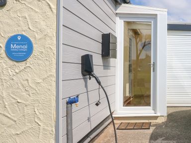 An exterior wall with a blue Menai Holiday Cottages sign an electric car charger and a glass door entrance at Flip Flops in Rhosneigr