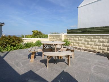 An outdoor patio area with a round wooden picnic table and benches surrounded by plants and a stone wall at Flip Flops in Rhosneigr