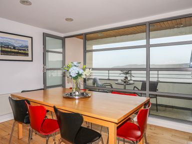 A dining room with wooden table and black and red chairs near glass doors overlooking a balcony with chairs and a table at Four The Quay in Red Wharf Bay