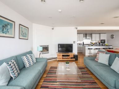 A living room with two blue sofas a clear glass coffee table a striped rug a television and a kitchen with bar stools at Four The Quay in Red Wharf Bay