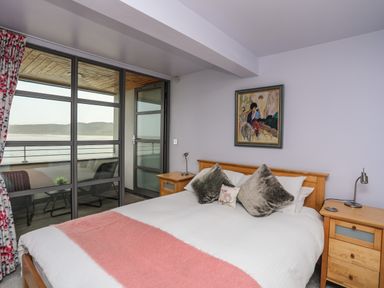 A bedroom with a wooden bed with pillows and a pink throw next to a balcony with chairs at Four The Quay in Red Wharf Bay