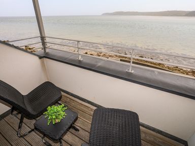 A balcony with two black wicker chairs and a small table with a plant overlooking a rocky beach and sea at Four The Quay in Red Wharf Bay
