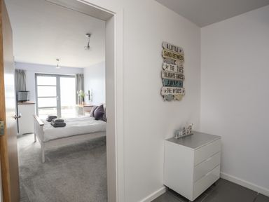 A bedroom with a bed and towels on it seen through a doorway with a small white chest of drawers and wall decor in the foreground at Four The Quay in Red Wharf Bay