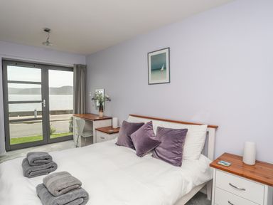 A bedroom with a double bed with purple pillows and folded towels next to a desk with a chair and a window overlooking water at Four The Quay in Red Wharf Bay