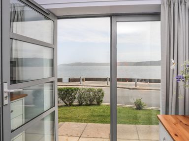 A view through open glass doors showing a small garden hedge a road and a body of water with hills in the background at Four The Quay in Red Wharf Bay