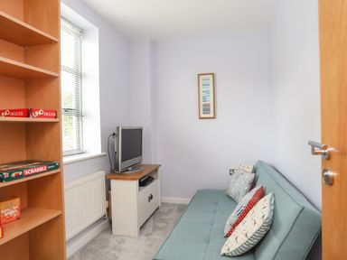 A small room with a sofa bed with pillows a TV on a stand and a wooden bookshelf with board games at Four The Quay in Red Wharf Bay