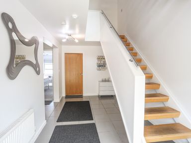 A hallway with wooden stairs metal handrail a wooden door two black rugs a white three drawer cabinet and a wavy wall mirror at Four The Quay in Red Wharf Bay