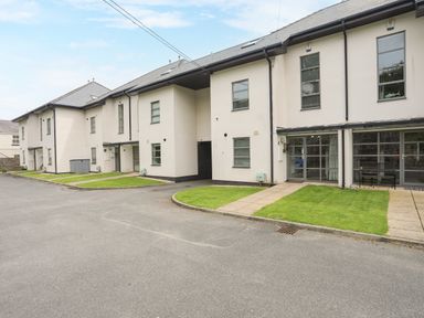 Exterior view of a two-story apartment building with white walls and multiple windows with small grass areas and paved walkways in front of each unit at Four The Quay in Red Wharf Bay