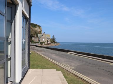 A coastal road alongside the sea with houses and a cliff under a blue sky at Four The Quay in Red Wharf Bay