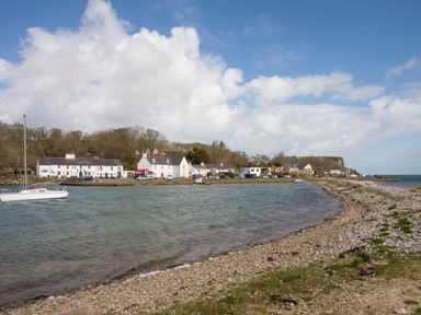 A rocky shoreline with water and white buildings along the coast with boats in the water at Four The Quay in Red Wharf Bay