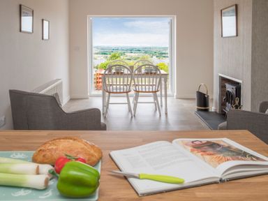 A dining area with white chairs and table near a large window overlooking a landscape with a kitchen counter in the foreground with bread vegetables and an open cookbook at Frondeg in Rhiwlas
