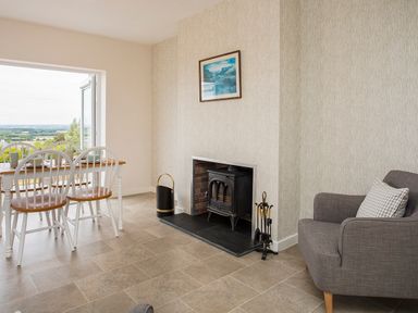 A dining area with a wooden table and white chairs a fireplace with a wood-burning stove and a gray armchair at Frondeg in Rhiwlas