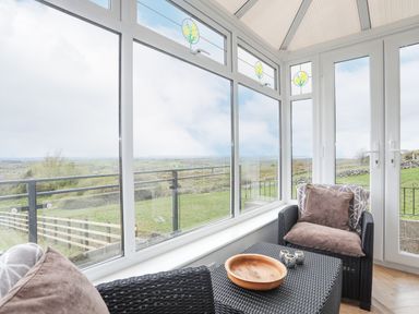 A sunroom with wicker chairs and a table overlooking a grassy landscape at Frondeg in Rhiwlas