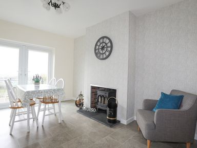 A dining area with a table and four chairs next to glass doors a fireplace with a clock above and a grey chair with a blue cushion at Frondeg in Rhiwlas