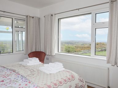 A bedroom with a bed covered in patterned bedding and folded towels by a window with curtains and a red chair at Frondeg in Rhiwlas