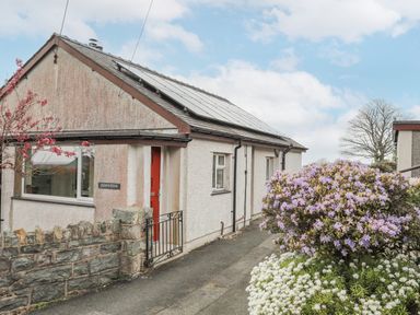 A small house with solar panels on the roof a red door a stone wall and flowering bushes in front at Frondeg in Rhiwlas