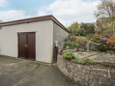 A small building with dark wooden double doors facing a paved area and a stone garden with plants and a metal water pump at Frondeg in Rhiwlas