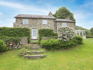 A stone cottage with a white door and windows surrounded by green bushes and a grassy lawn at Fron Fawr in Nefyn