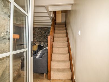 A narrow carpeted staircase with wooden railing next to a living area with grey chairs and stone wall at Fron Fawr in Nefyn