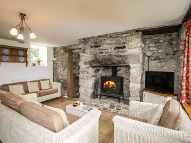 A living room with stone walls a fireplace with a wood stove wicker sofas a television and a wooden shelf at Fron Fawr in Nefyn