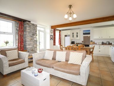 A living room and kitchen with wicker sofa and armchair a dining table and white kitchen cabinets at Fron Fawr in Nefyn