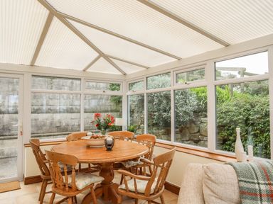 A dining area with a round wooden table and six chairs in a glass conservatory with plants outside at Fron Fawr in Nefyn